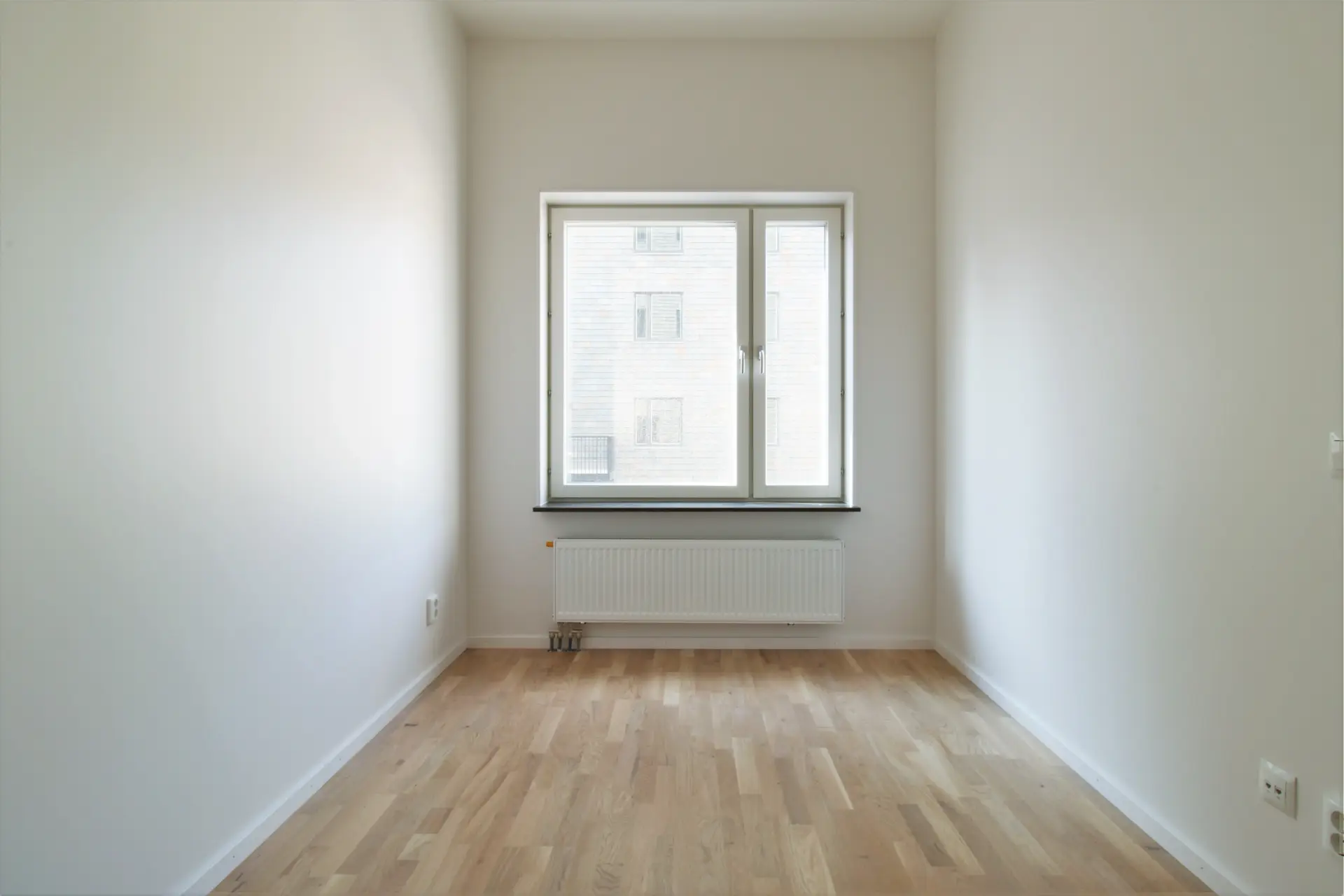 Tomt rum med vita väggar, trägolv och ett stort fönster som vetter mot en byggnad och visar naturligt ljus. Ett element finns under fönstret.