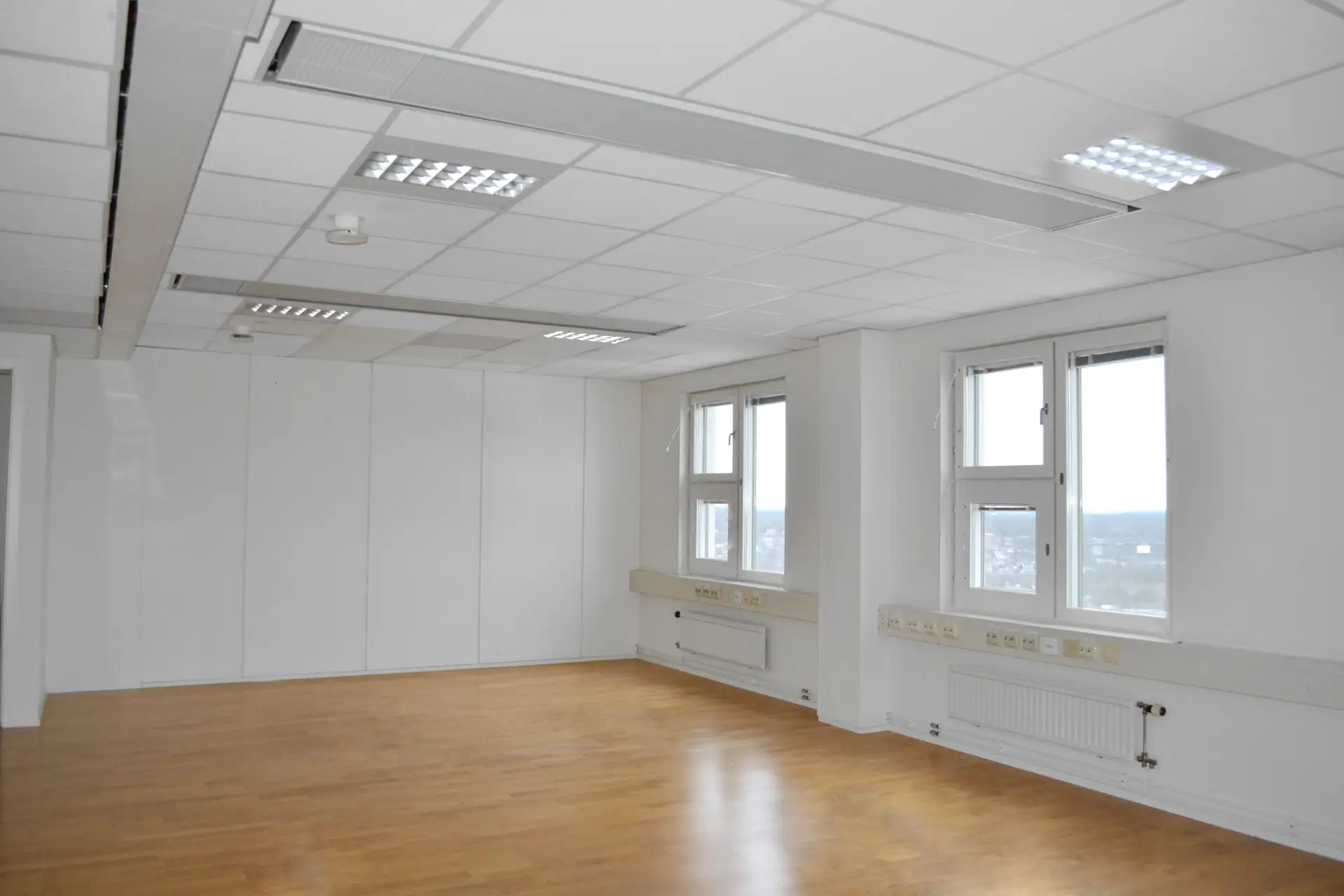 Ett tomt kontorsrum med vita väggar, stora fönster som släpper in naturligt ljus och ett trägolv.