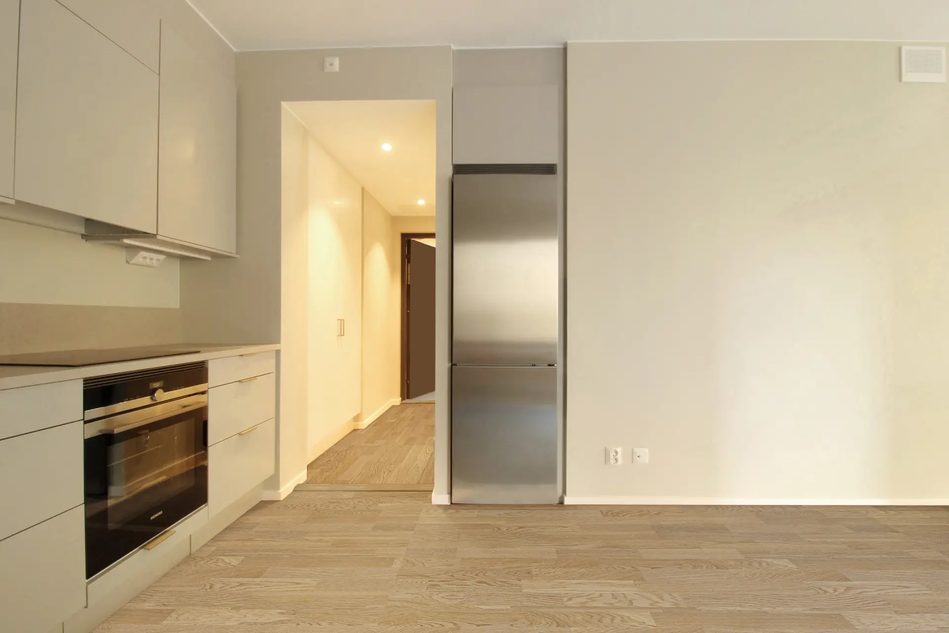 Ett kök med ljusa trägolv, vita väggar och inbyggda rostfria vitvaror inklusive ugn och kylskåp.