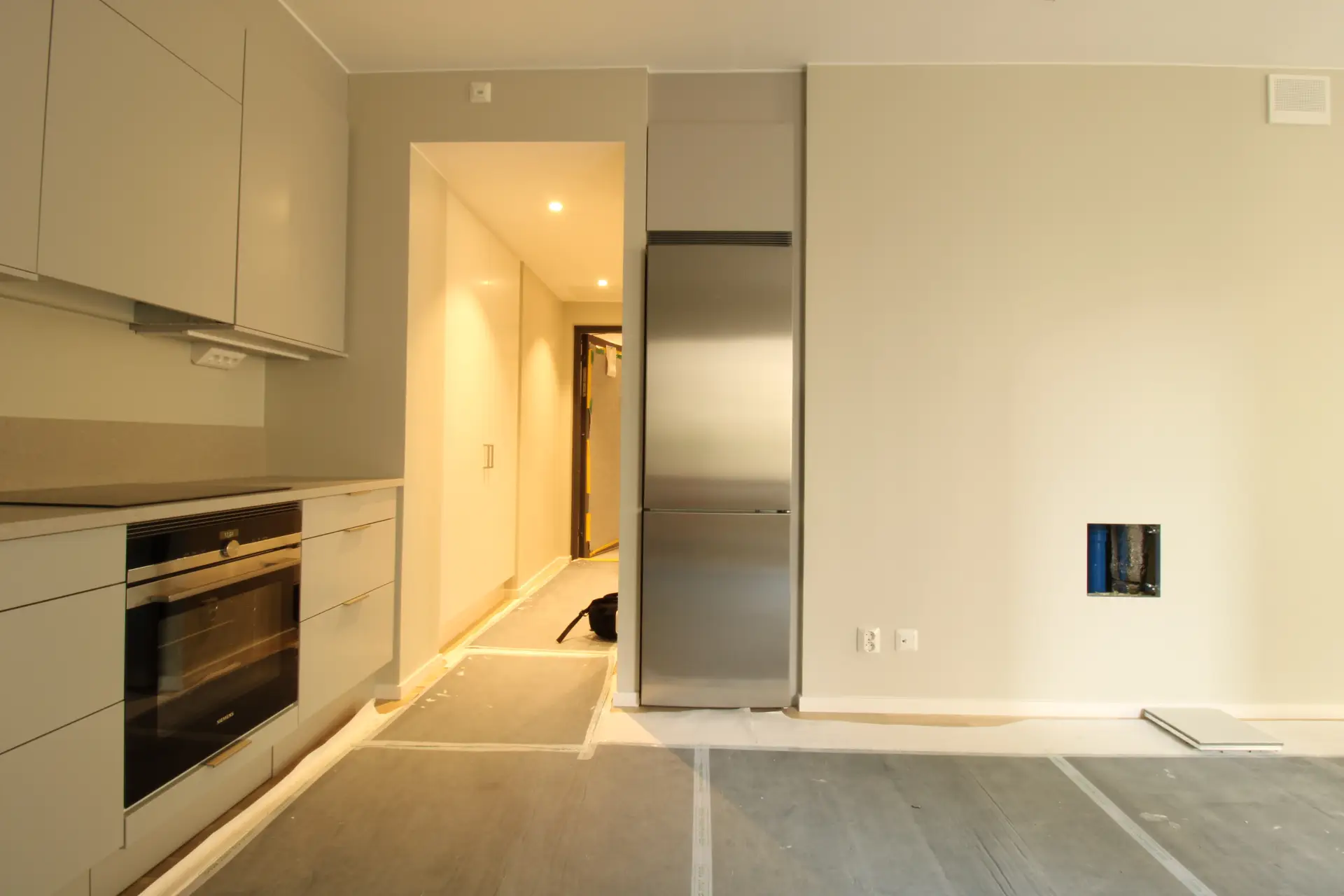 Ett kök under uppbyggnad med ljusgråa luckor, en ugn och ett kylskåp, med skyddande överdrag på golvet.