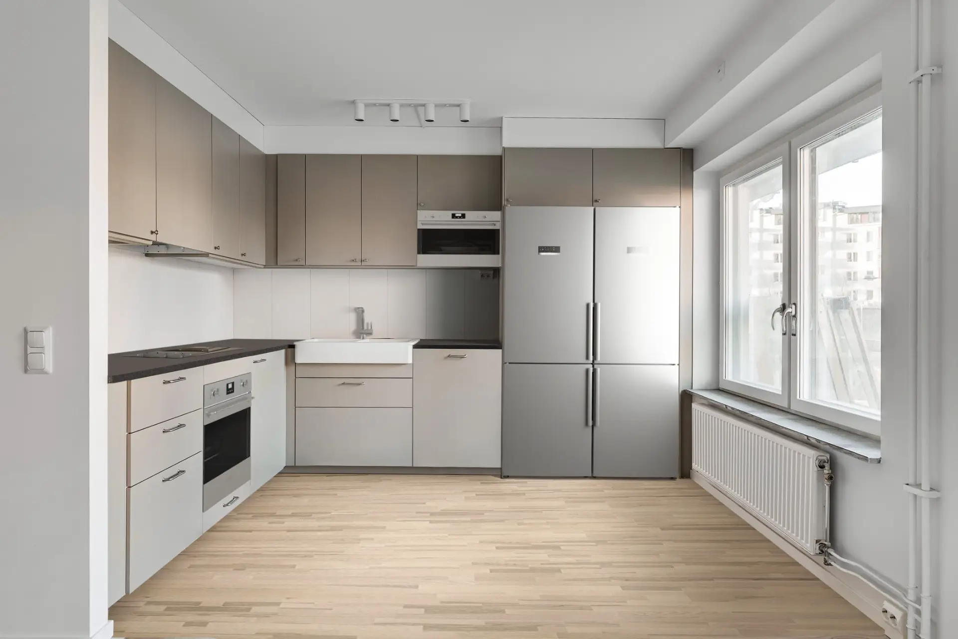 Modernt kök med ljusa trägolv, beige köksluckor och rostfria vitvaror inklusive kylskåp, ugn och integrerad diskmaskin. Naturligt ljus fyller rummet genom stora fönster.