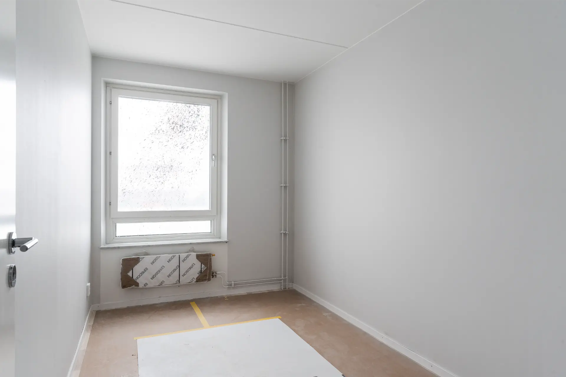 Ett minimalistiskt tomt rum med ett smutsigt fönster, ljusgrå väggar och ett emballerat element under fönstret. golvet är delvis täckt med en skyddsduk.