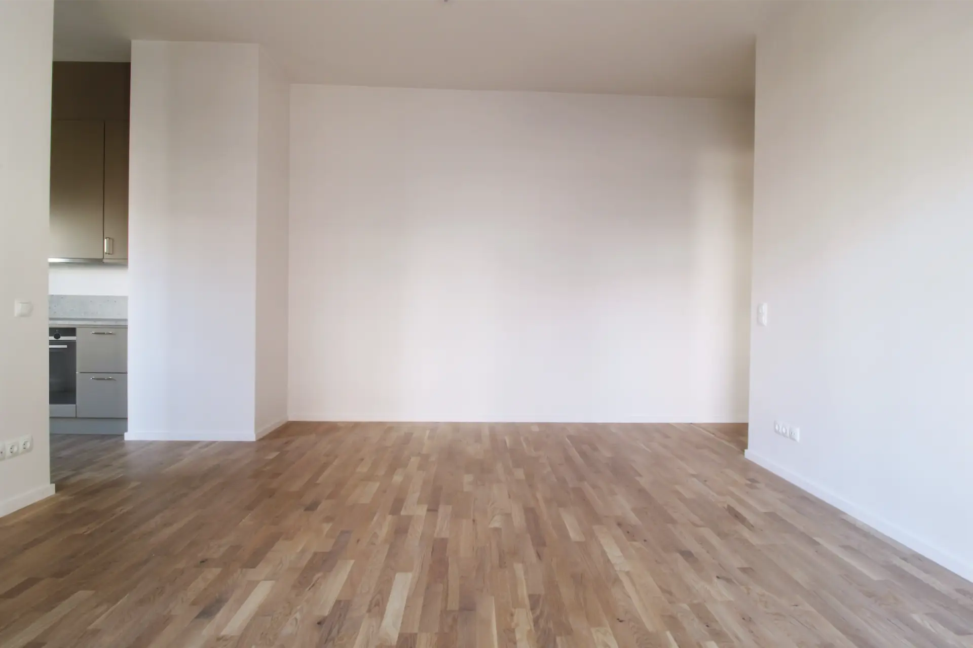 Ett rymligt, tomt rum med ljusa väggar, trägolv och en liten köksdel synlig i det vänstra hörnet med gråa skåp och vitvaror.